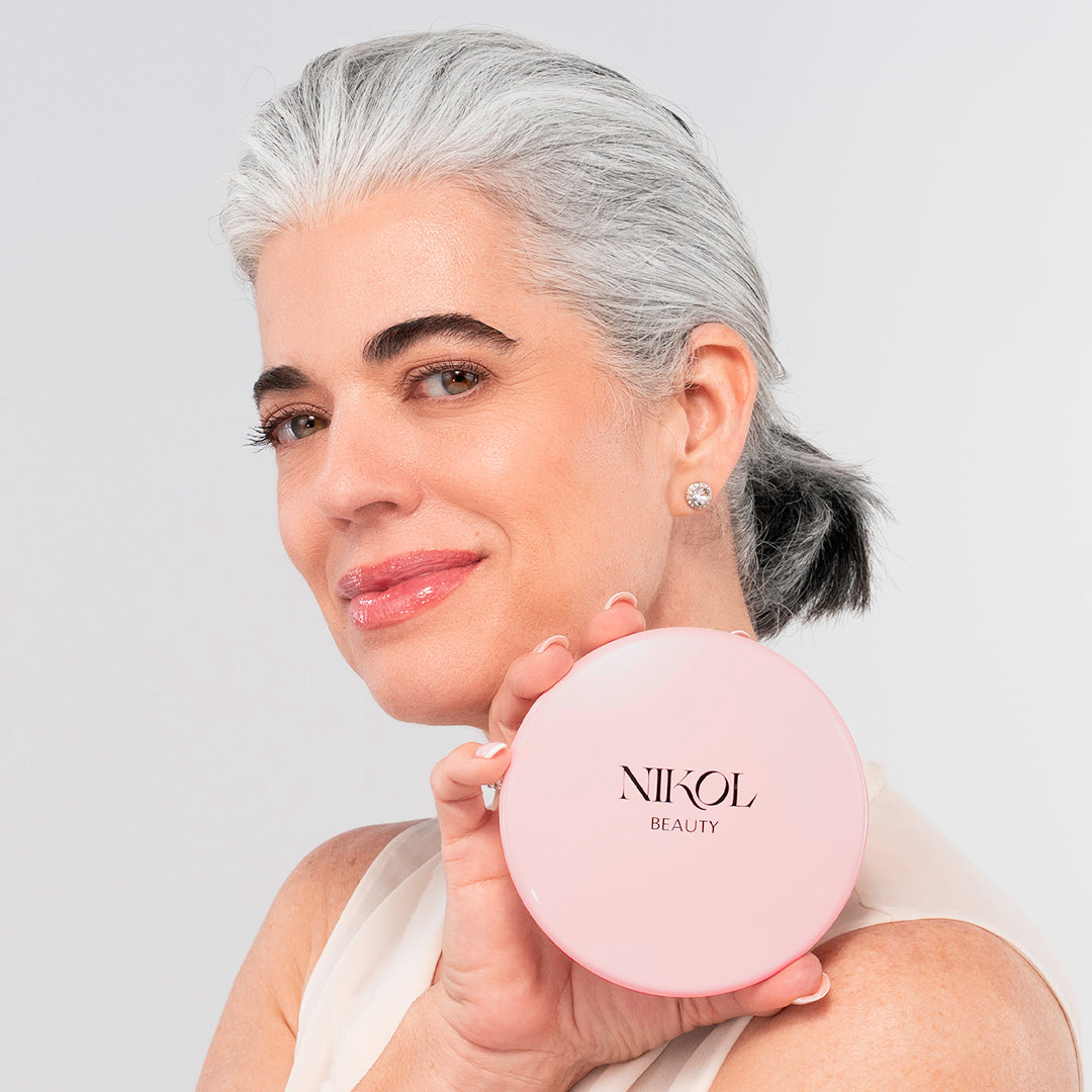 A woman with gray hair pulled back smiles and holds the Nikol Beauty 10x Magnification Mirror near her face, as if admiring herself. She wears a sleeveless top and stud earrings, set against a light background.