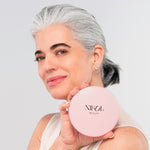 A woman with gray hair pulled back smiles and holds the Nikol Beauty 10x Magnification Mirror near her face, as if admiring herself. She wears a sleeveless top and stud earrings, set against a light background.