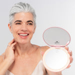 A smiling woman with short gray hair holds the Nikol Beauty 10x Magnification Mirror, an LED ring light makeup mirror. She wears a beige camisole and small earrings against a neutral background.