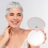 A smiling woman with short gray hair holds the Nikol Beauty 10x Magnification Mirror, an LED ring light makeup mirror. She wears a beige camisole and small earrings against a neutral background.