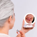 A woman with gray hair applies lip gloss while looking into the round, illuminated Nikol Beauty 10x Magnification Mirror, which reflects her smiling lips.