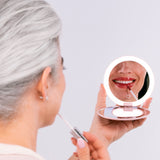 A woman with gray hair applies lip gloss while looking into the round, illuminated Nikol Beauty 10x Magnification Mirror, which reflects her smiling lips.