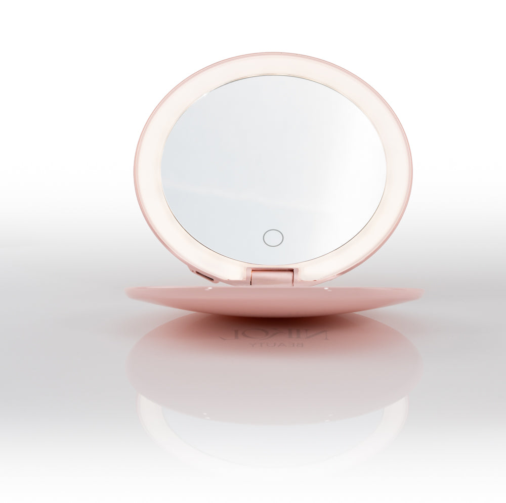 The Nikol Beauty 10x Magnification Mirror is a round, pink makeup mirror with a built-in LED ring light, shown open and standing upright on a reflective white surface.