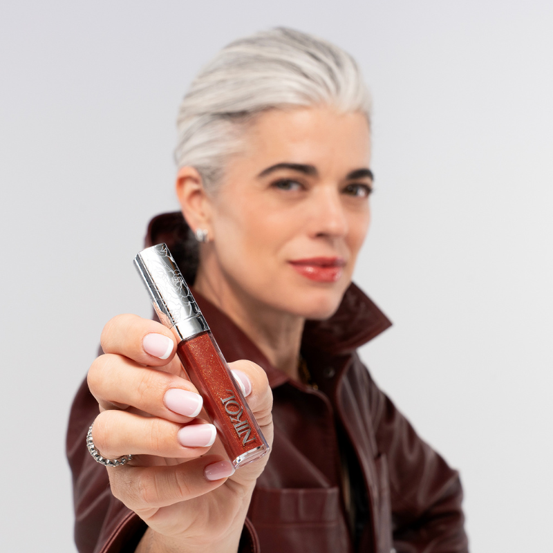 A mature woman with short gray hair and a maroon leather jacket smiles softly as she holds up Nikol Beauty’s IGNITE LIP GLOSS #176, drawing focus to the shimmering gloss in her hand.