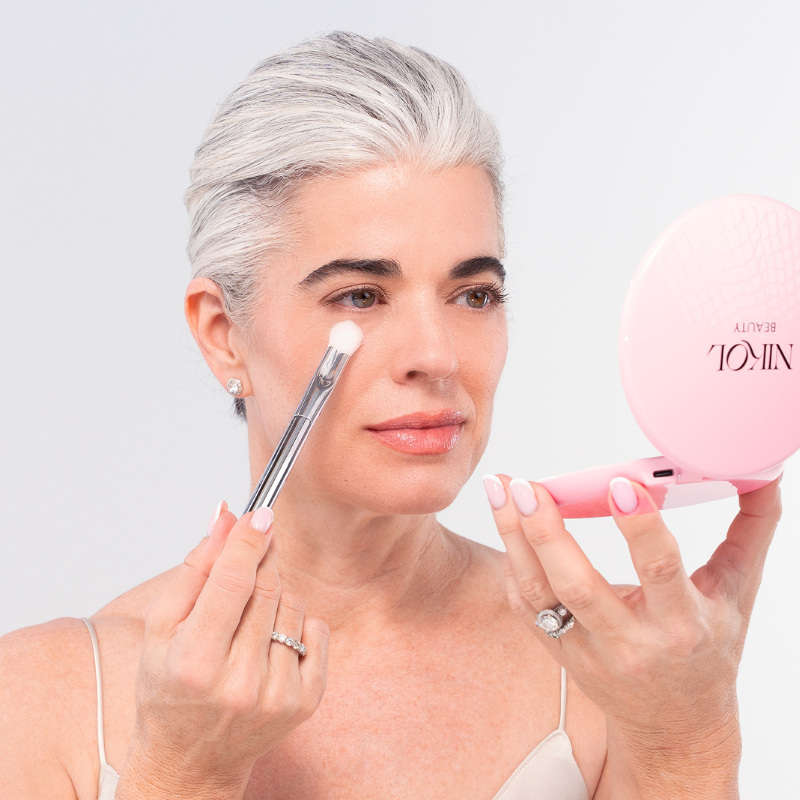 A woman with gray hair applies makeup using the Nikol Beauty SOFT SET TECHNIQUE™ BRUSH while holding a pink compact mirror. Wearing a light-colored top and rings, she looks focused on her reflection.