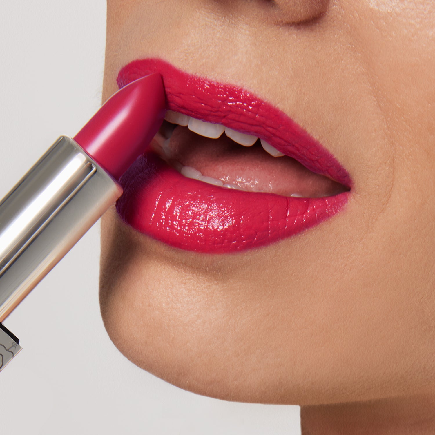 A close-up of someone applying Nikol Beauty’s Fleur Shiny Velvet Cream Lipstick #34, a vibrant pink vegan formula, showing smooth, glossy lips, white teeth, and part of the metallic lipstick tube.
