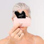 A woman with short gray hair holds a black Gua Sha Stainless Steel tool from Nikol Beauty in a pale pink case, partially covering her face. She wears a thin strap top and silver ring, set against a plain white background.