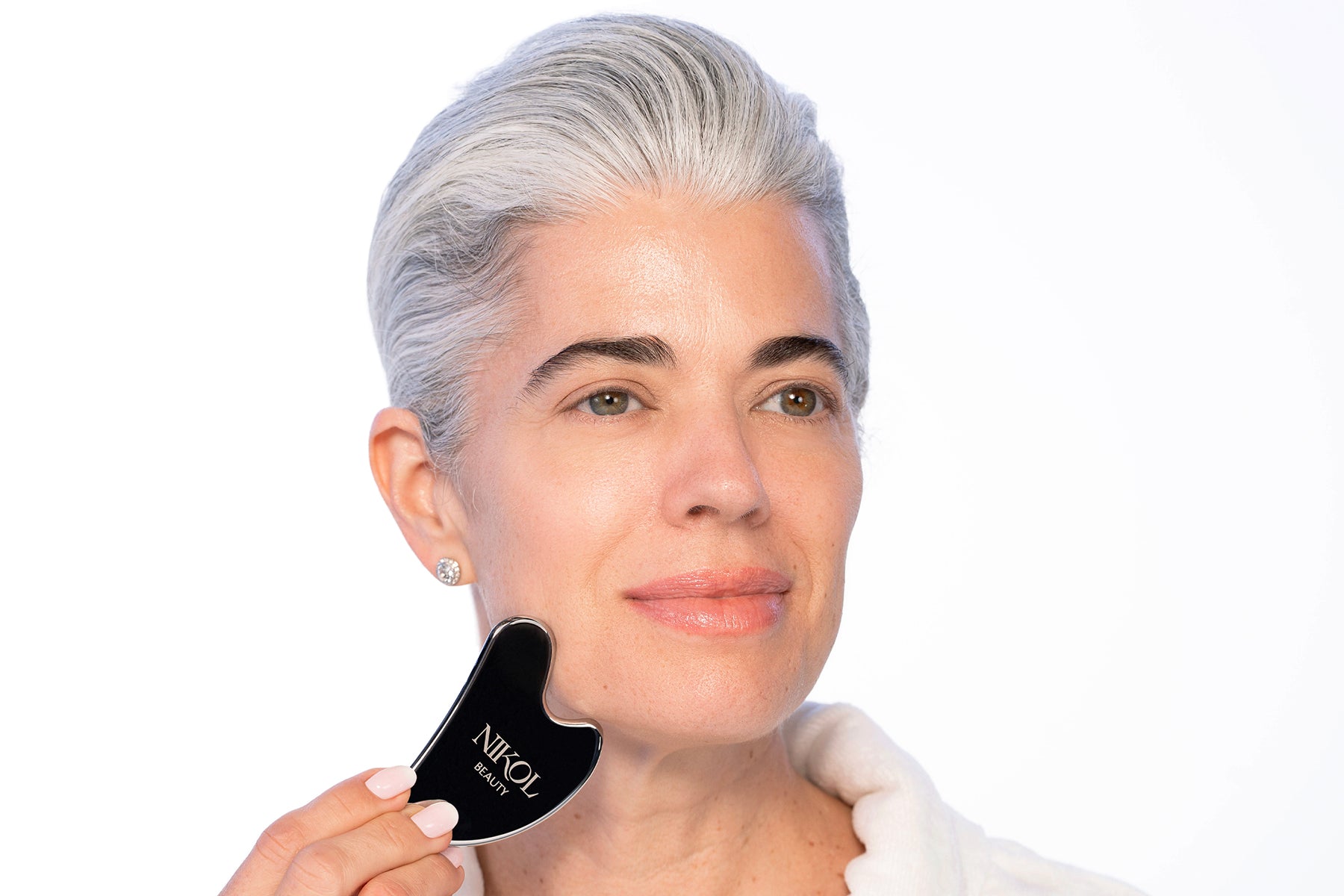 A woman with short gray hair uses the Nikol Beauty Gua Sha Stainless Steel tool on her cheek, wearing a white robe and small stud earrings. She looks calm and relaxed, enjoying lymphatic drainage benefits against a plain background.