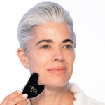A woman with short gray hair smiles softly as she uses the Nikol Beauty Gua Sha Stainless Steel tool on her cheek, promoting lymphatic drainage. She wears a white top and stud earrings against a light background.