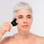 A woman with short gray hair uses the Nikol Beauty Gua Sha Stainless Steel facial massage tool on her cheek. She wears a light camisole and appears calm, set against a plain, light background.
