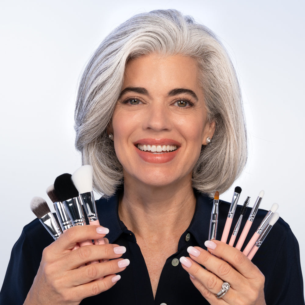 A smiling woman with gray hair, dressed in a dark top and showing off her manicured nails, holds the Nikol Beauty Essential Makeup Brush Set in both hands against a plain, light background.