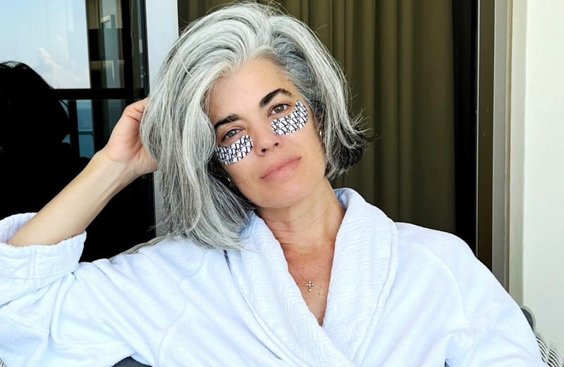 A woman with shoulder-length gray hair wearing a white robe and patterned under-eye patches sits relaxed, resting her head on one hand. She is seated indoors near a window or door with beige curtains in the background.