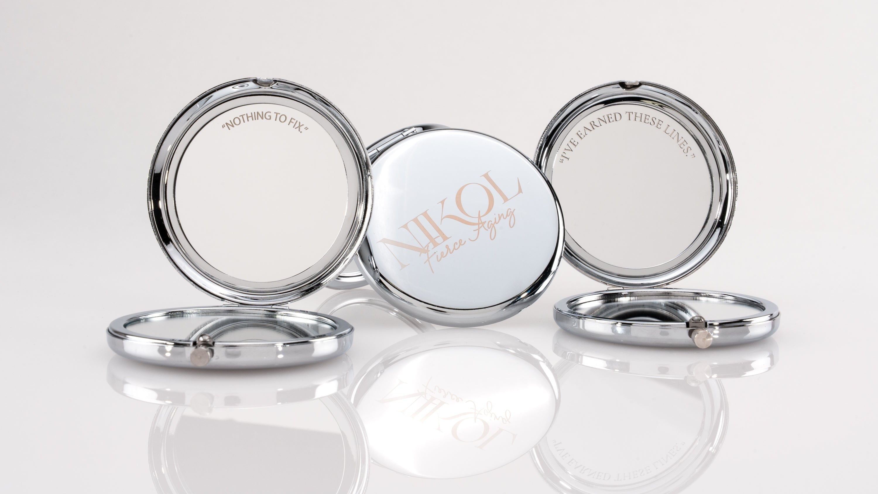 Three round, silver Fierce Aging Mini Compact Mirrors from Nikol Beauty stand open—one reads NIKOL Faster Aging, while the sides say NOTHING TO FIX and I’VE EARNED THESE LINES!, celebrating confidence and portable beauty.
