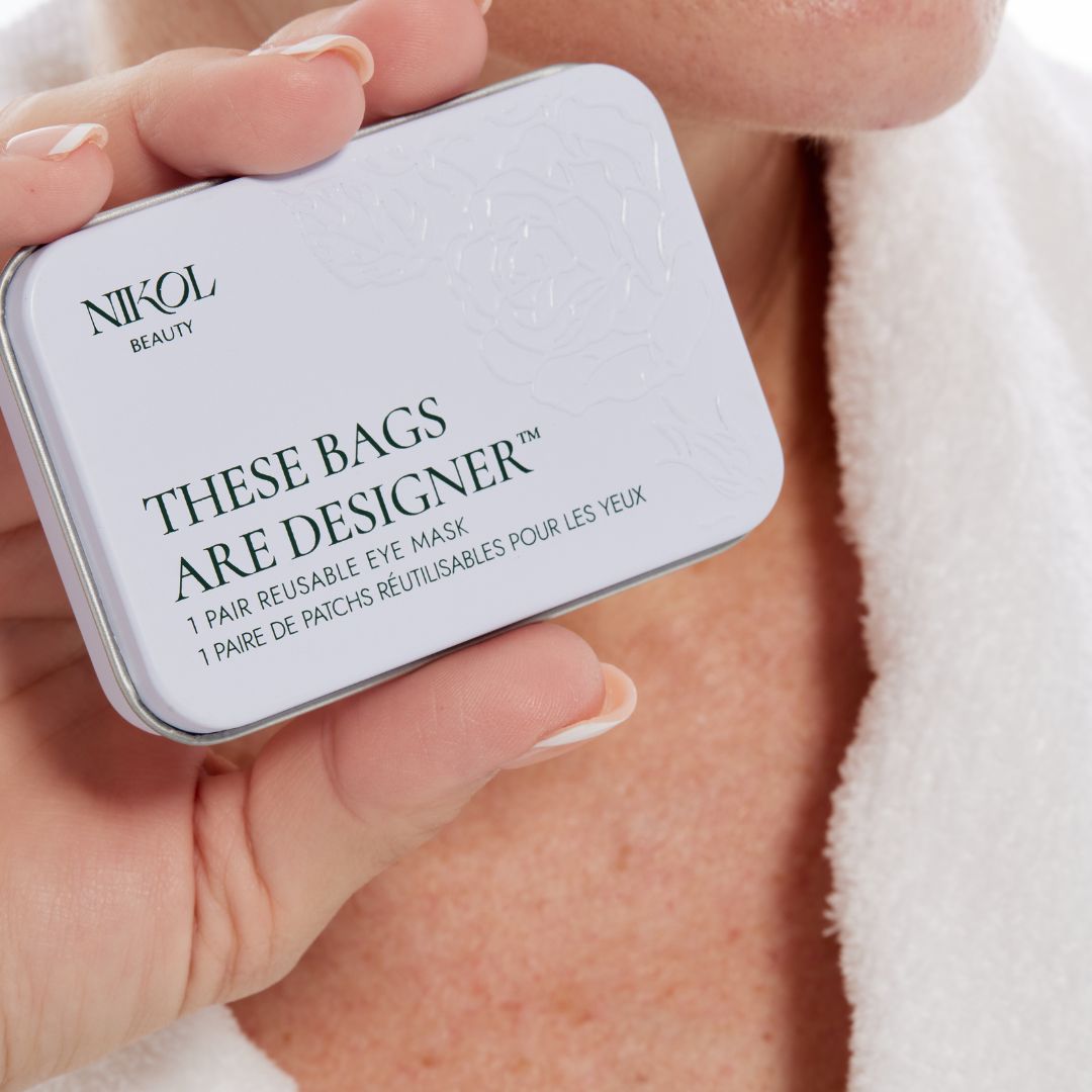 A hand holding the closed rose-embossed These Bags Are Designer tin beside a white robe