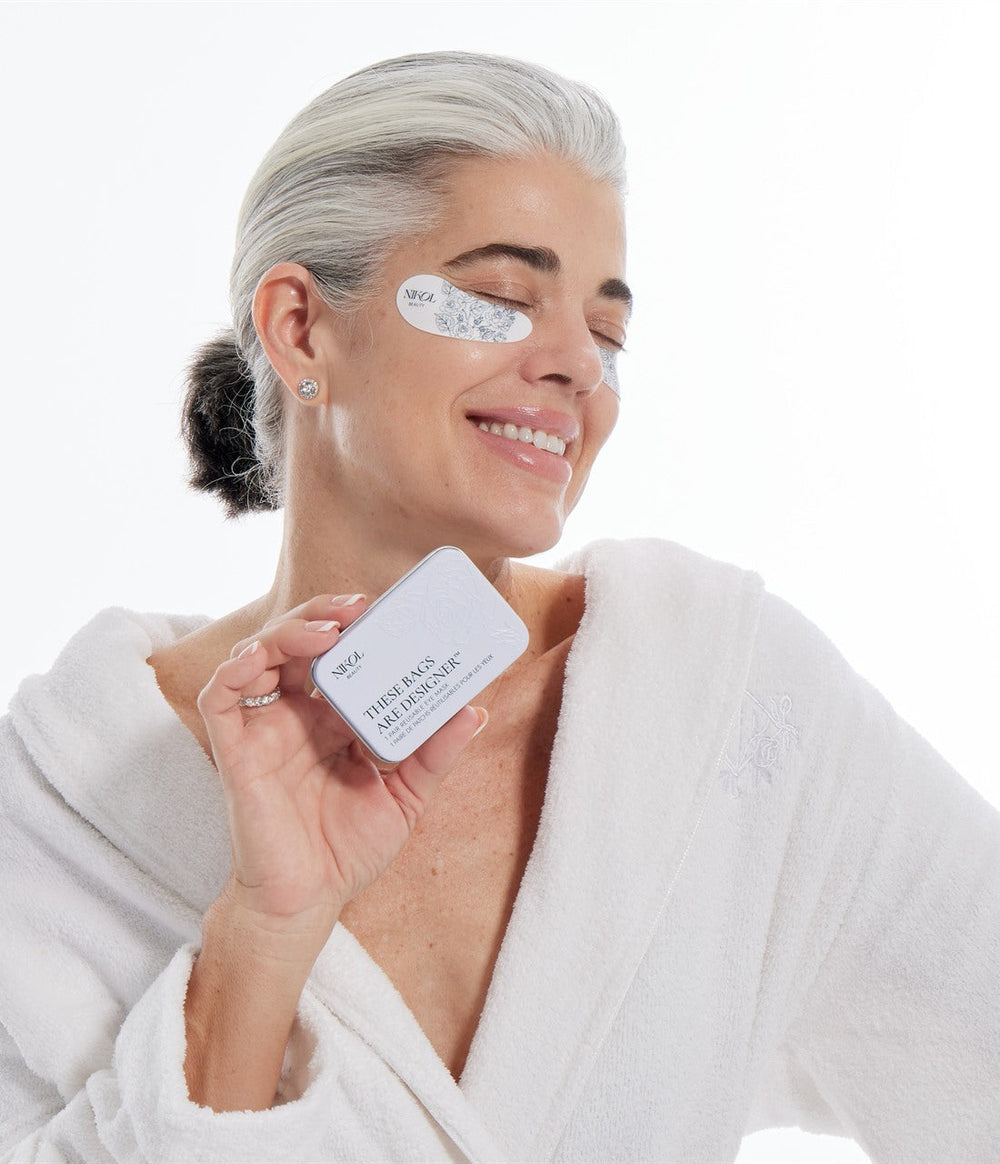 Nikol in a white robe smiling with a These Bags Are Designer eye mask under one eye, holding the tin