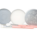 Two round silver tins—one with a textured pad and the other with Lux Brush Soap by Nikol Beauty—are displayed open. In front, two pink-handled Nikol Beauty makeup brushes rest on a white background.