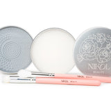 Two round silver tins—one with a textured pad and the other with Lux Brush Soap by Nikol Beauty—are displayed open. In front, two pink-handled Nikol Beauty makeup brushes rest on a white background.