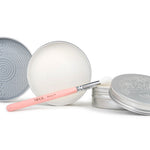 Three silver tins of Nikol Beauty Lux Brush Soap—one open to display the cleansing surface, one showing the soap, and a pink makeup brush with silver ferrule in front—an all-natural cleaner for effortlessly clean tools.