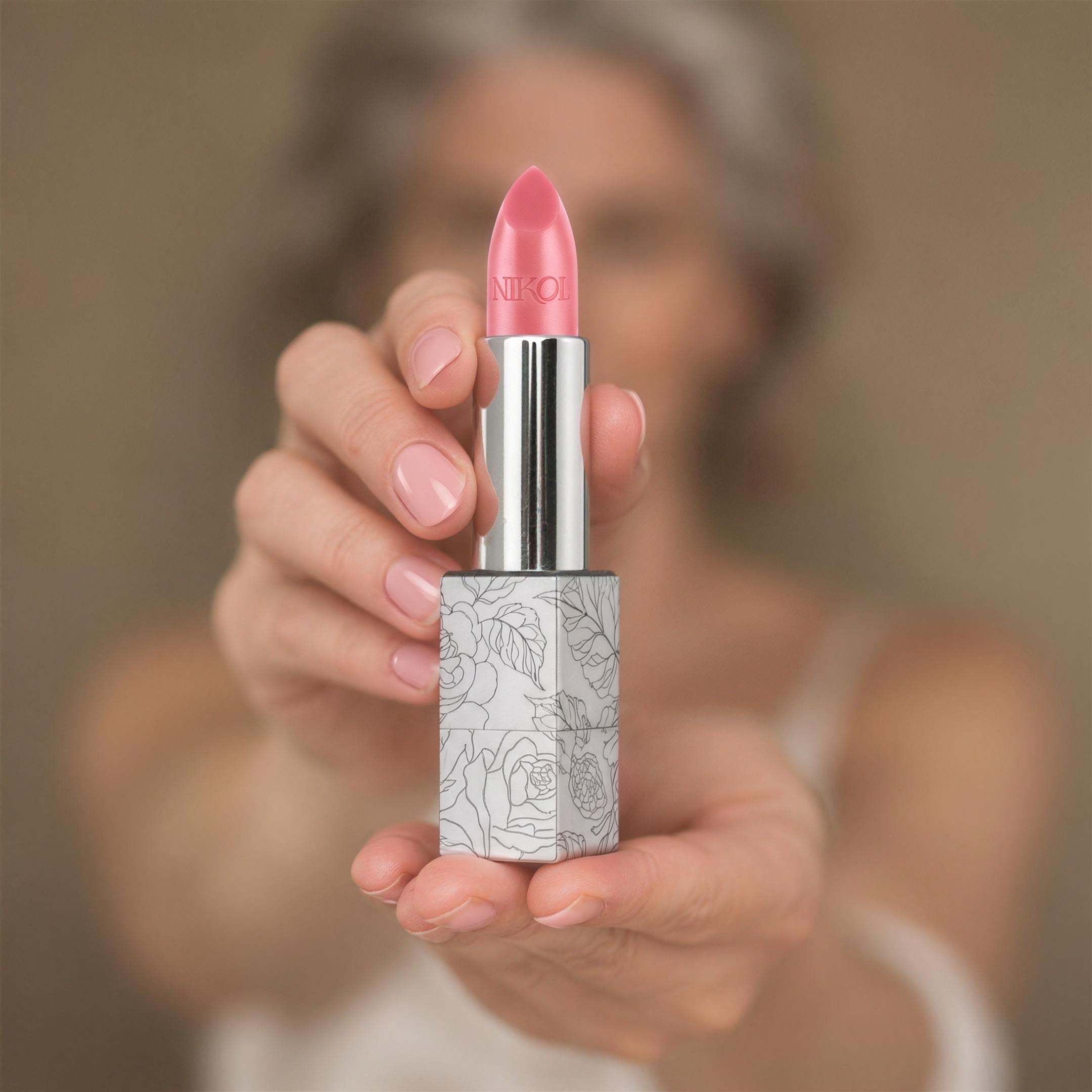 The uncapped Rose Lip Balm, a rose-pink bullet embossed with NIKOL in its rose-engraved silver case, held out toward the camera in two manicured hands, Nikol softly blurred behind
