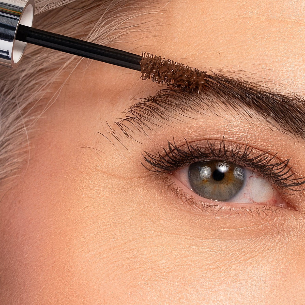 Close-up of the Brow Mascara wand brushing soft brown color through a mature brow, the open silver tube just in frame