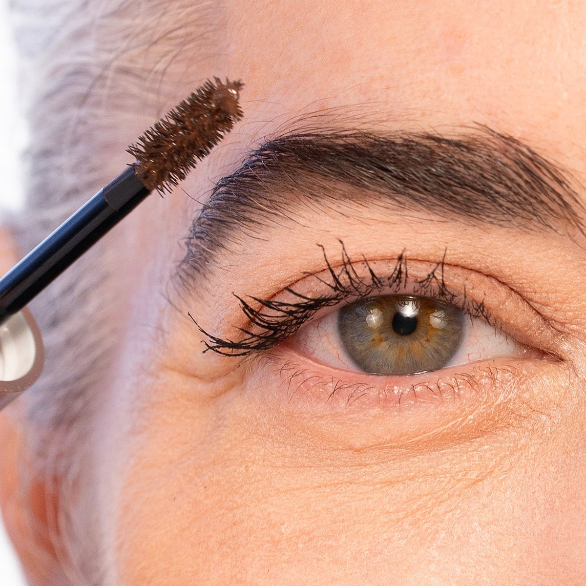 The Brow Mascara wand, loaded with brown color, lifted toward a defined mature brow beside the open tube