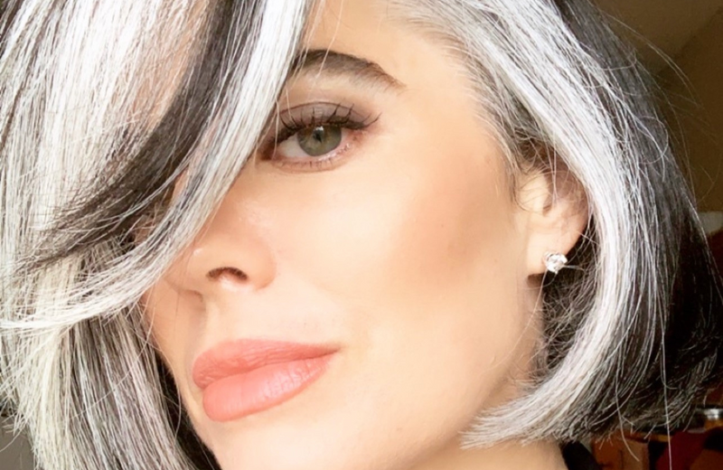 A woman with a stylish short bob haircut featuring black and white streaks, wearing subtle makeup and a stud earring, looks confidently at the camera.