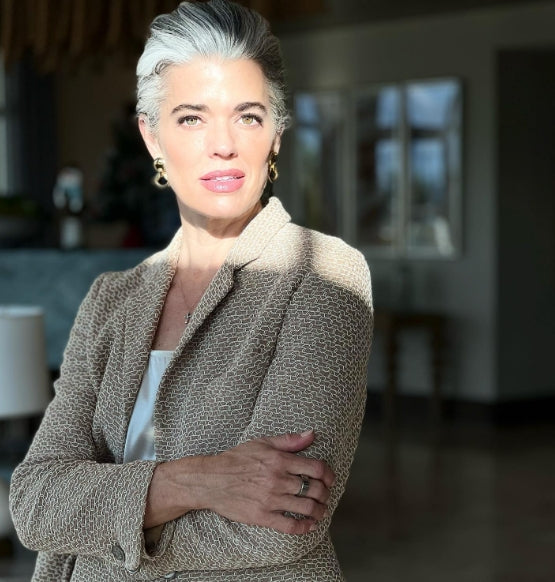 A woman with short gray hair and a beige patterned blazer stands indoors with arms crossed, looking confidently at the camera in natural sunlight.