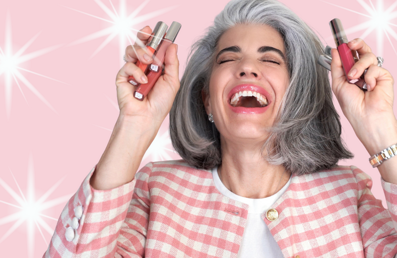 A joyful woman with gray hair and a pink plaid blazer holds up lip gloss tubes, smiling widely against a pink background with white starbursts.