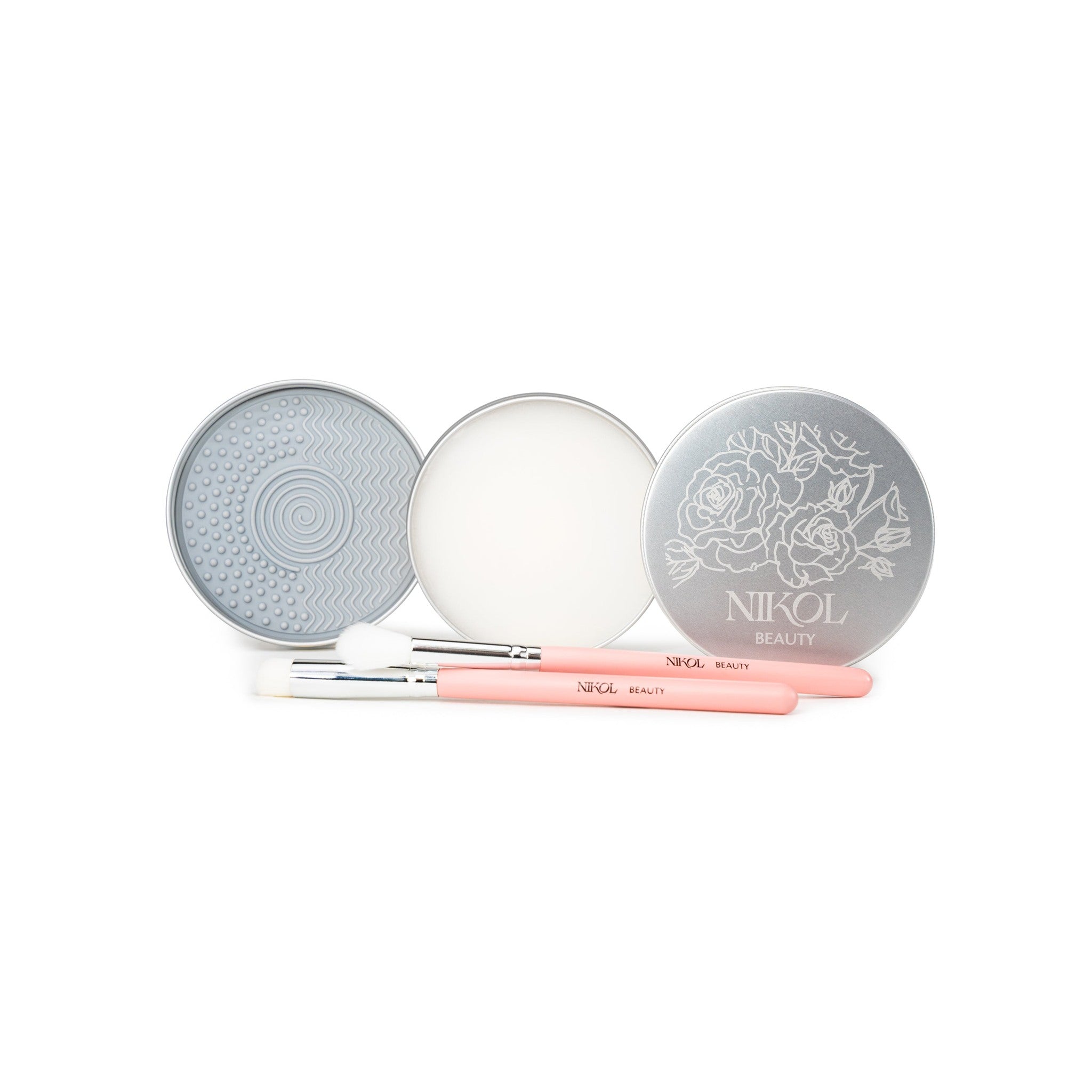 Two round silver tins—one with a textured pad and the other with Lux Brush Soap by Nikol Beauty—are displayed open. In front, two pink-handled Nikol Beauty makeup brushes rest on a white background.