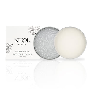 A cylindrical tin with a textured geometric lid holds a round bar of Lux Brush Soap by Nikol Beauty next to its white packaging adorned with floral line art. Infused with coconut oil, the soap gently cleans makeup brushes.