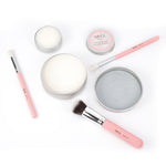 A flat lay of Nikol Beauty Lux Brush Soap, pink-handled brushes, tins of vegan brush cleaner, coconut oil brush soap, and a round cleaning pad, all neatly arranged on a white background.