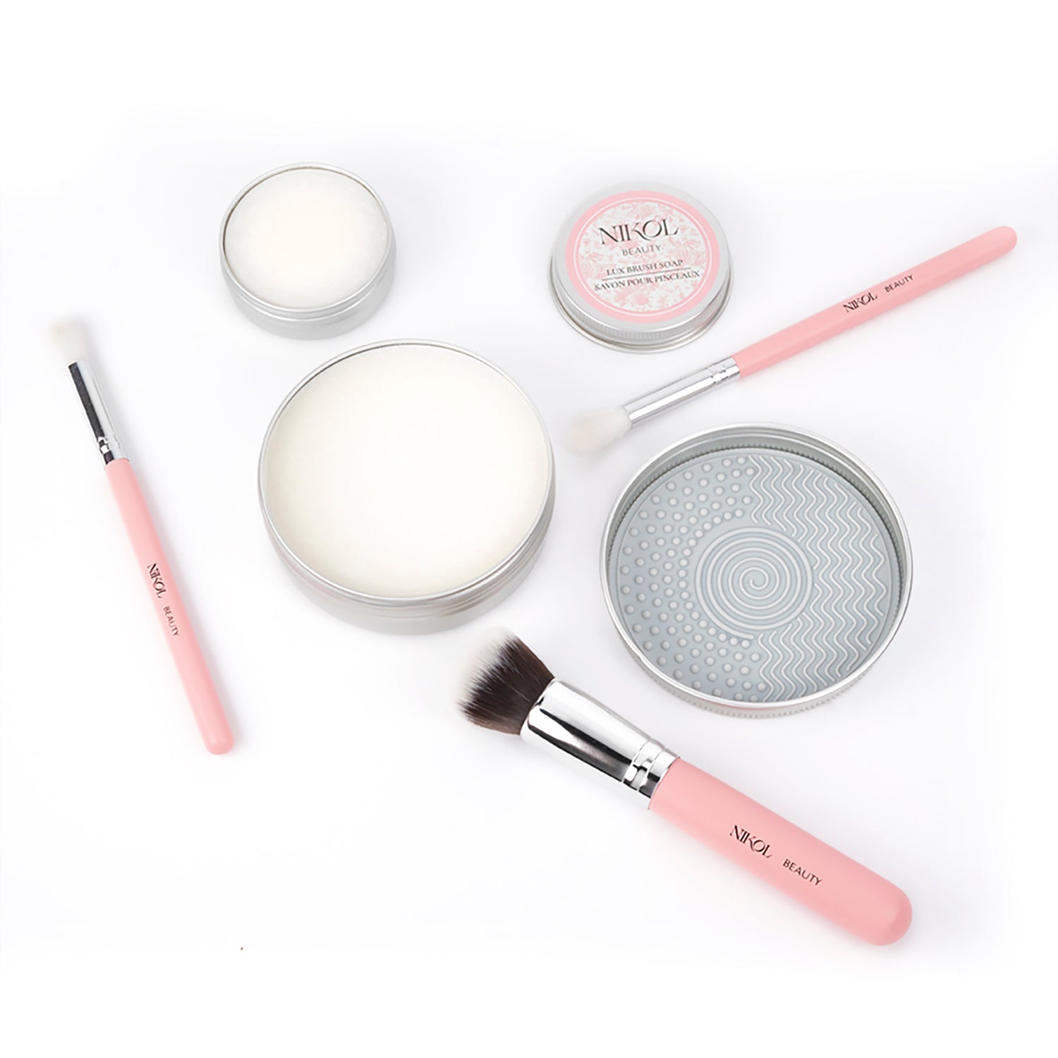 A flat lay of Nikol Beauty Lux Brush Soap, pink-handled brushes, tins of vegan brush cleaner, coconut oil brush soap, and a round cleaning pad, all neatly arranged on a white background.