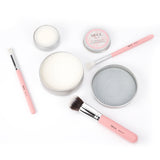 A flat lay of Nikol Beauty Lux Brush Soap, pink-handled brushes, tins of vegan brush cleaner, coconut oil brush soap, and a round cleaning pad, all neatly arranged on a white background.