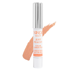 A tube of Nikol Beautys Just Peachy Color Corrector, created for undereye discoloration, stands against a white background, with a peach-colored swipe behind it. The tube shows metallic accents and the products name and weight.