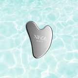 A Gua Sha Stainless Steel tool from Nikol Beauty, engraved with the brands name, rests against a backdrop of calm, light blue rippling water, designed to boost lymphatic drainage.