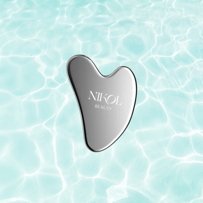 A Gua Sha Stainless Steel tool from Nikol Beauty, engraved with the brands name, rests against a backdrop of calm, light blue rippling water, designed to boost lymphatic drainage.